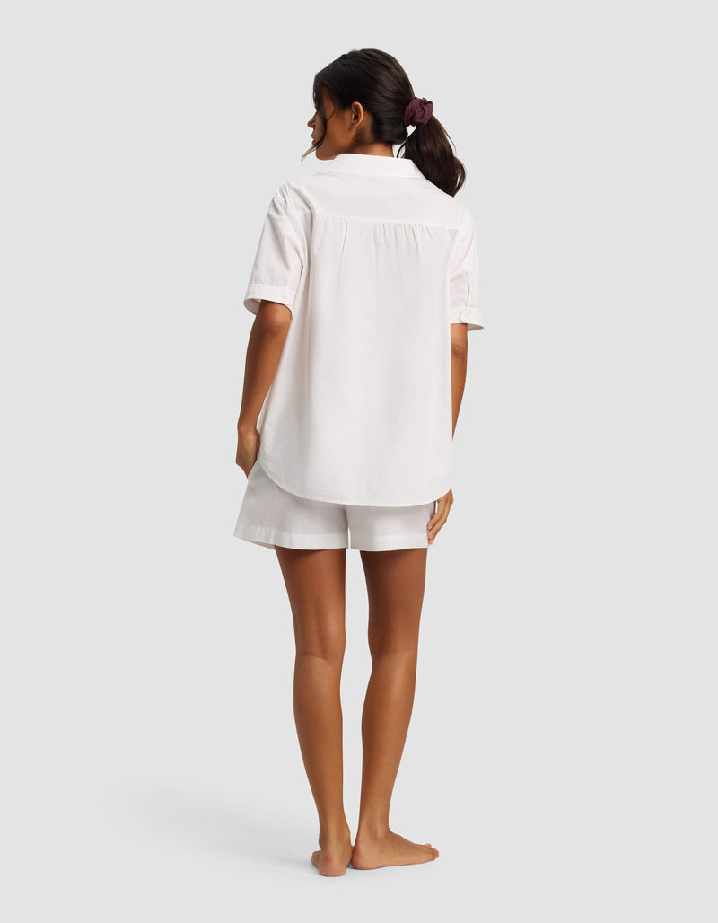 A woman with her dark hair tied back in a scrunchie stands barefoot, facing away, wearing the Cozy Earth Women's Sutton Short Sleeve Pajama Set in white. The background is plain and light gray.
