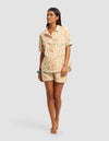 A woman stands barefoot in the Cozy Earth Women's Sutton Short Sleeve Pajama Set, featuring a matching button-up shirt and shorts with a pastel floral pattern, against a plain light gray background.|Color:Wildflower
