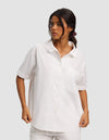 A woman stands against a plain light gray background wearing the Cozy Earth Women's Sutton Short Sleeve Pajama Top in white, paired with matching white shorts.|Color:White
