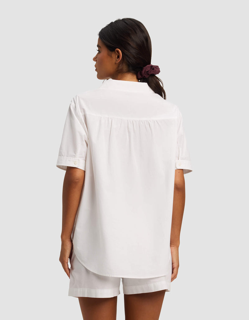 A woman with dark hair in a ponytail, tied with a dark scrunchie, is shown from the back wearing the Cozy Earth Women's Sutton Short Sleeve Pajama Top and matching white shorts against a plain light gray background.