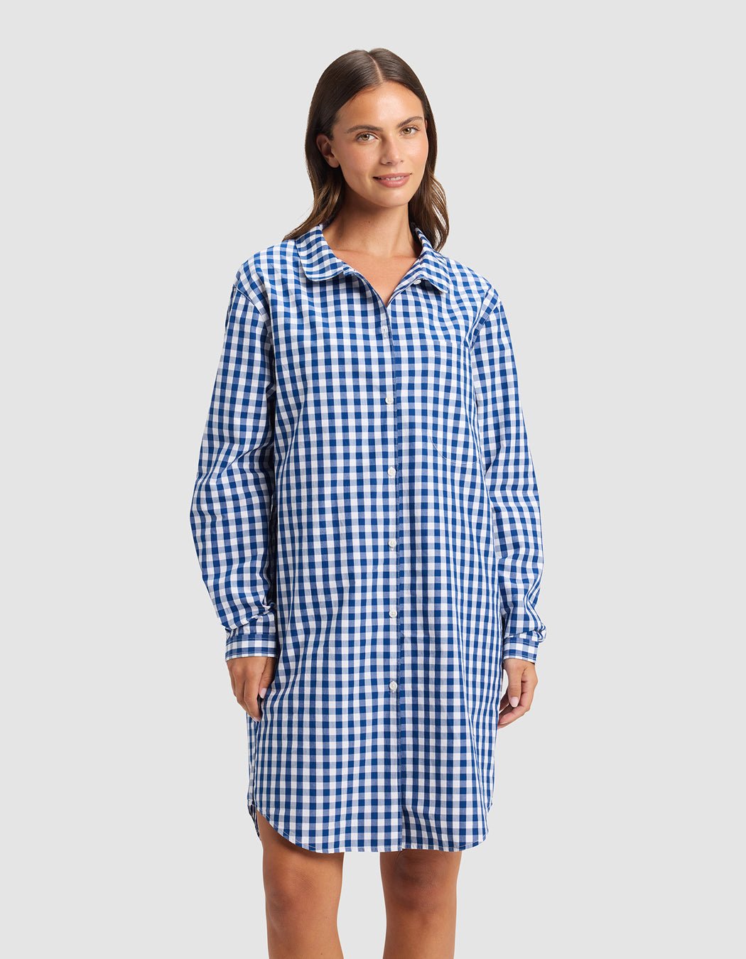 A woman with straight brown hair wears the Cozy Earth Women's Sutton Sleep Dress, a blue and white checkered, long-sleeve shirt dress that falls above the knees. She smiles slightly against a plain light background.
