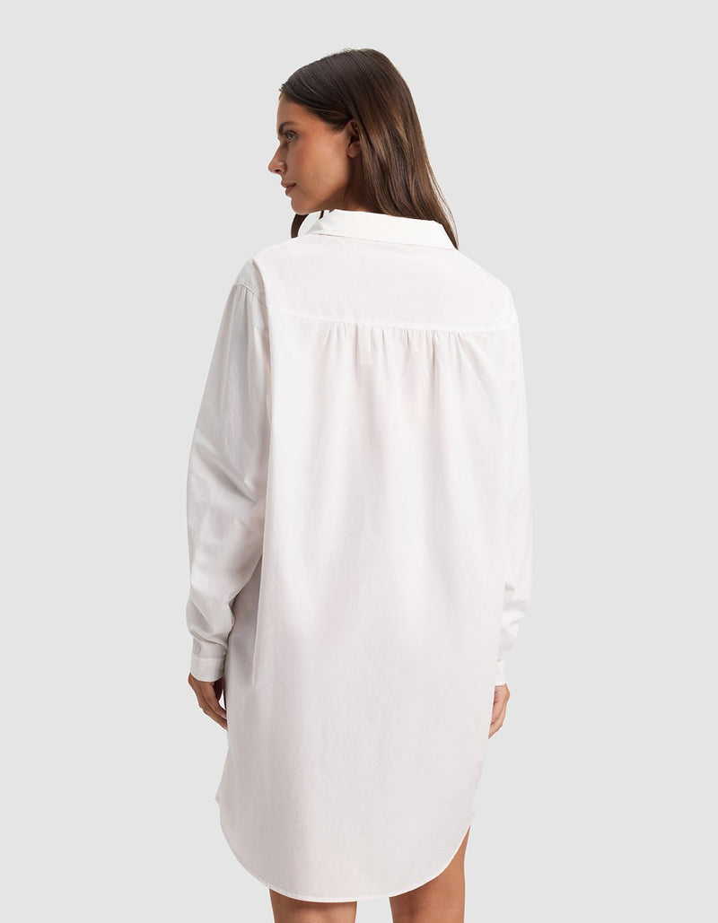 A woman with long brown hair faces away, wearing the Cozy Earth Women's Sutton Sleep Dress—a loose-fitting white, long-sleeve shirt dress—set against a plain light gray background.