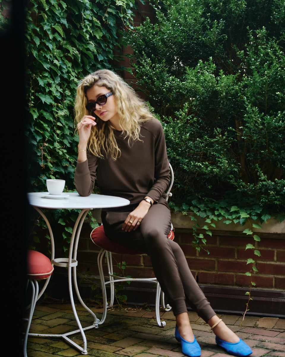 A woman wearing a brown Cozy Earth Brushed Bamboo Pullover and Jogger set, styled with black sunglasses and holding a white mug, standing outdoors in front of a green hedge. |Color:Espresso
