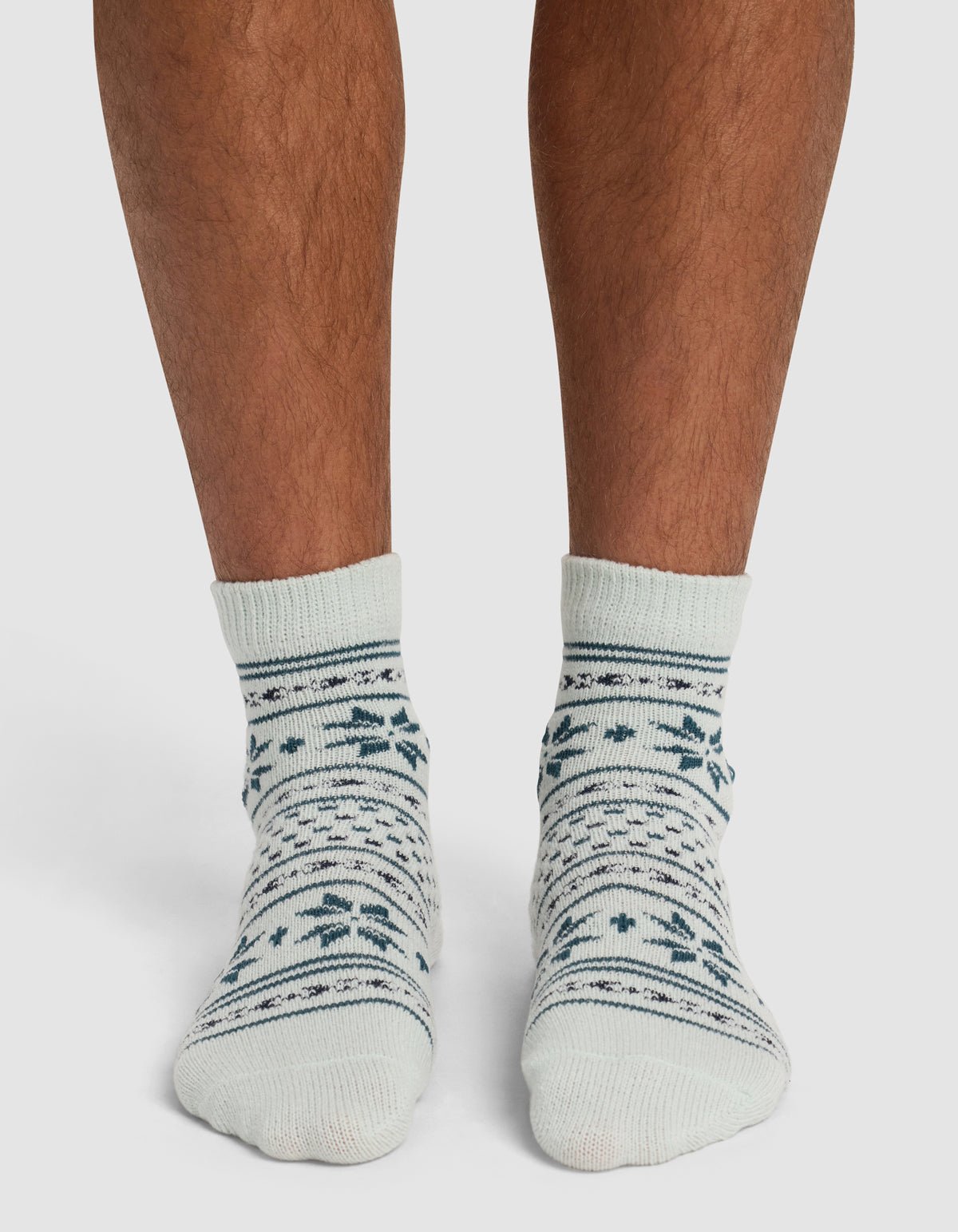 A person wearing Cozy Earth's The Snowflake Plush Modern Crew Sock—cream-colored with dark green and blue snowflake and stripe patterns—stands on a white surface.|Color:Mist