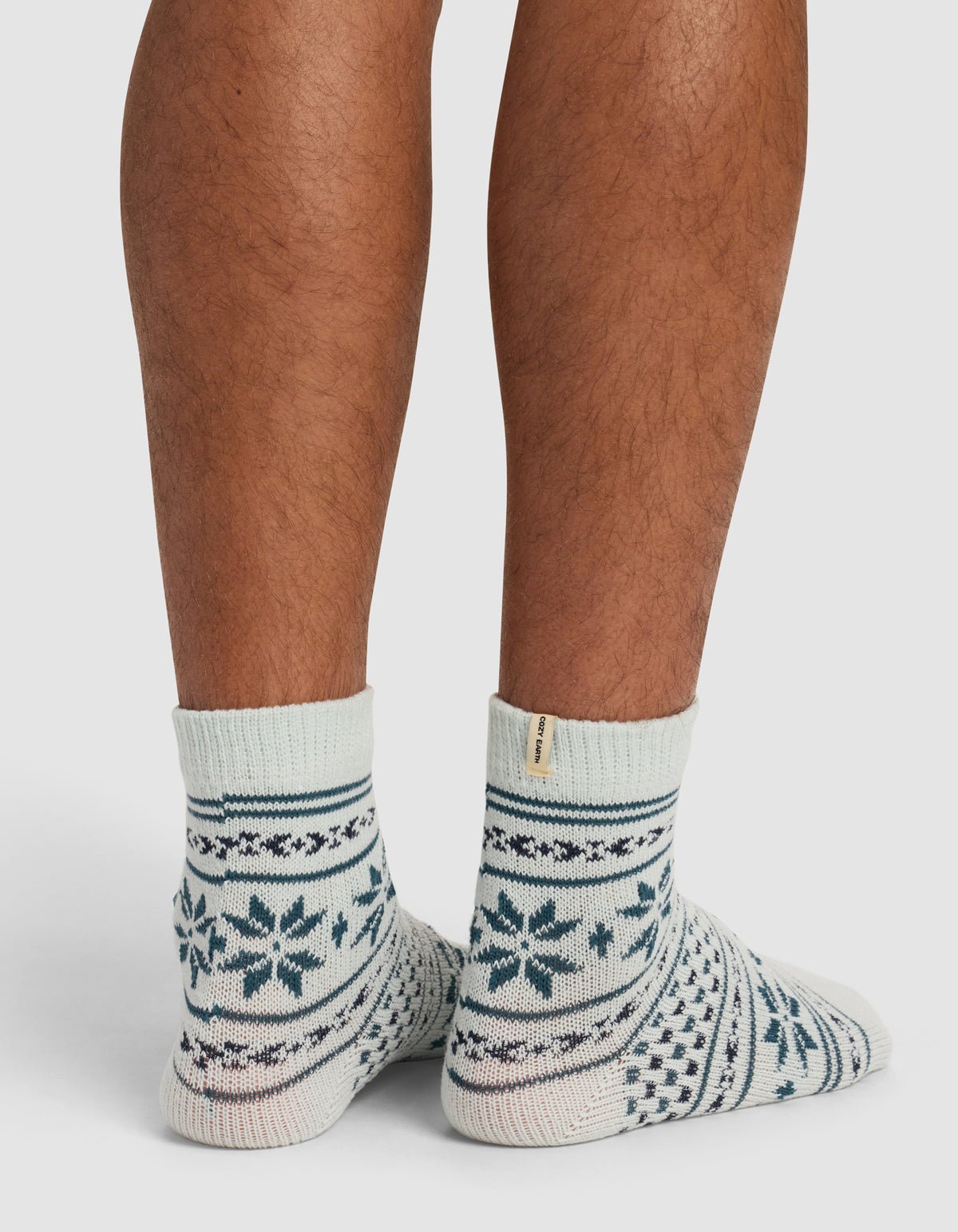 A person’s lower legs and feet are shown standing on a white surface, wearing Cozy Earth’s The Snowflake Plush Modern Crew Sock—light-colored socks with dark blue snowflakes and geometric patterns.|Color:Mist