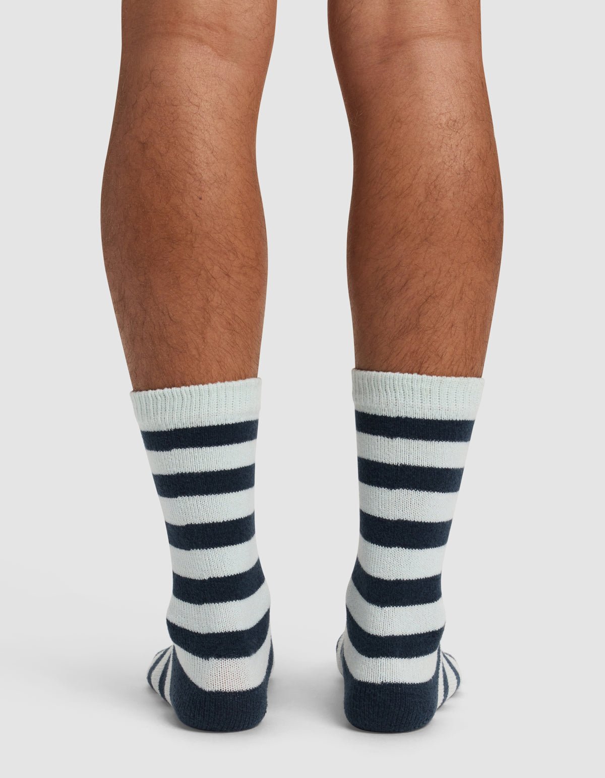 Back view of a person's lower legs wearing Cozy Earth's The Striped Plush Lounge Sock, featuring thick navy blue horizontal stripes on white, standing on a white background.|Color:Mist Stripe