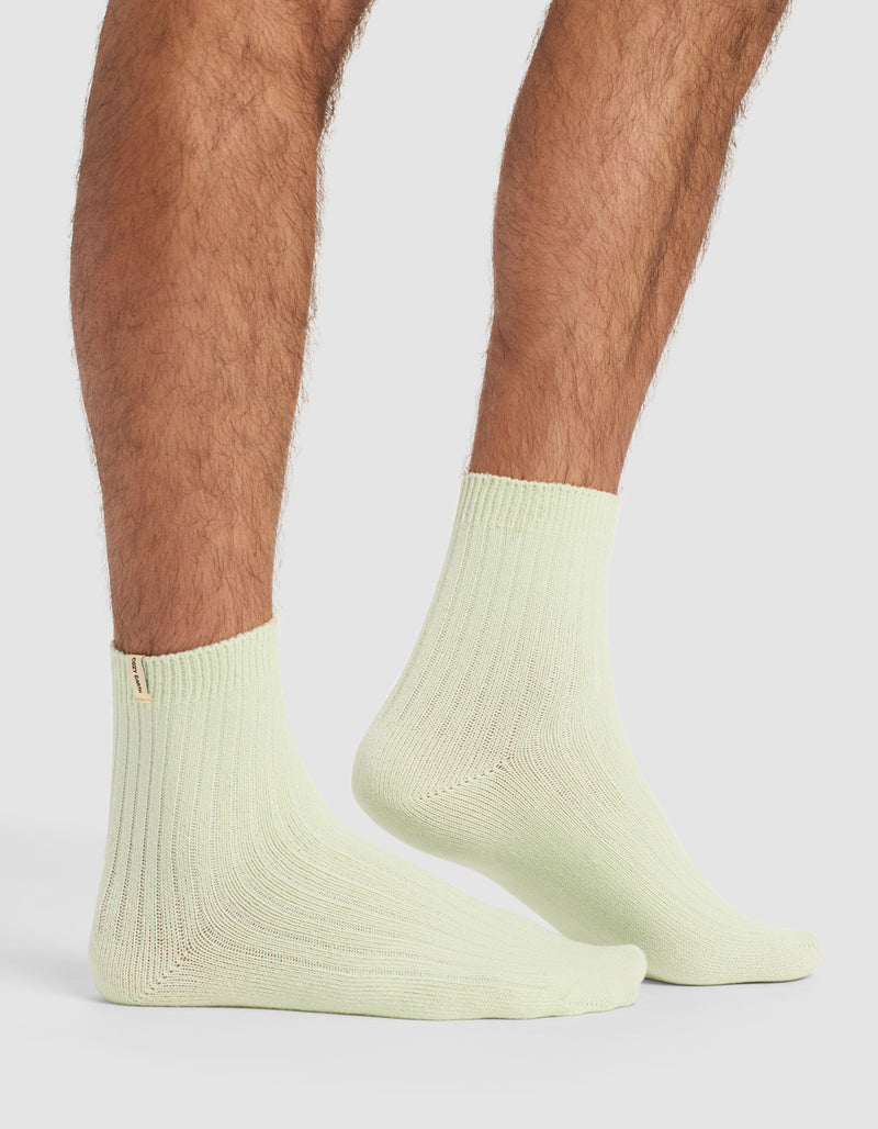 A person with hairy legs stands on a white surface wearing light green socks from Cozy Earth's Plush Lounge Quarter Sock 3-Pack.