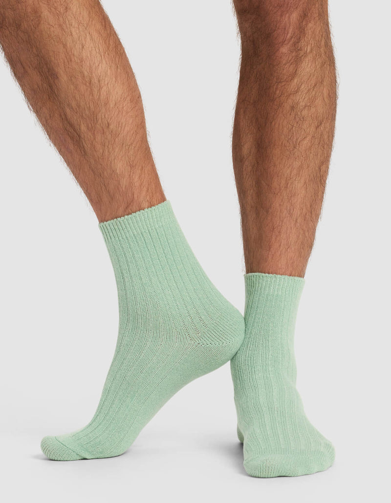 A person with hairy legs wears Cozy Earth's Men's Plush Lounge Quarter Sock 3-Pack in light green, standing with one foot slightly behind the other against a plain white background.