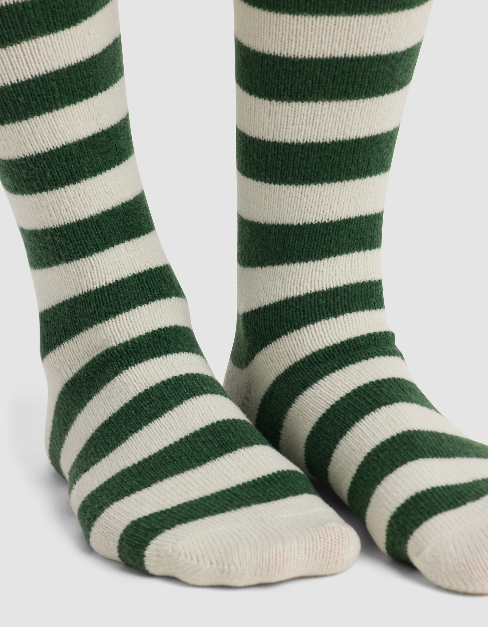 Close-up of feet wearing Cozy Earth's The Striped Plush Lounge Sock, featuring thick, horizontal dark green stripes on white, standing on a white background.|Color:Celadon Stripe
