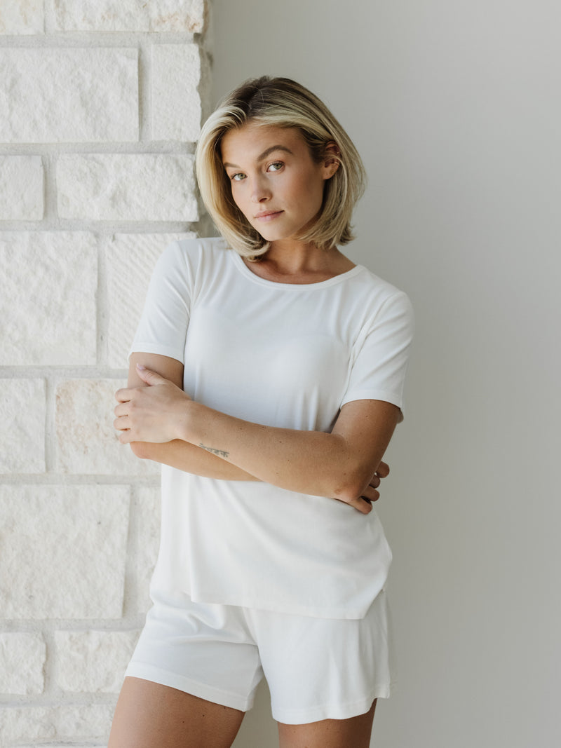 Woman wearing a white Cozy Earth Rib Knit Short Sleeve Lounge Top and white shorts against a white stone background
