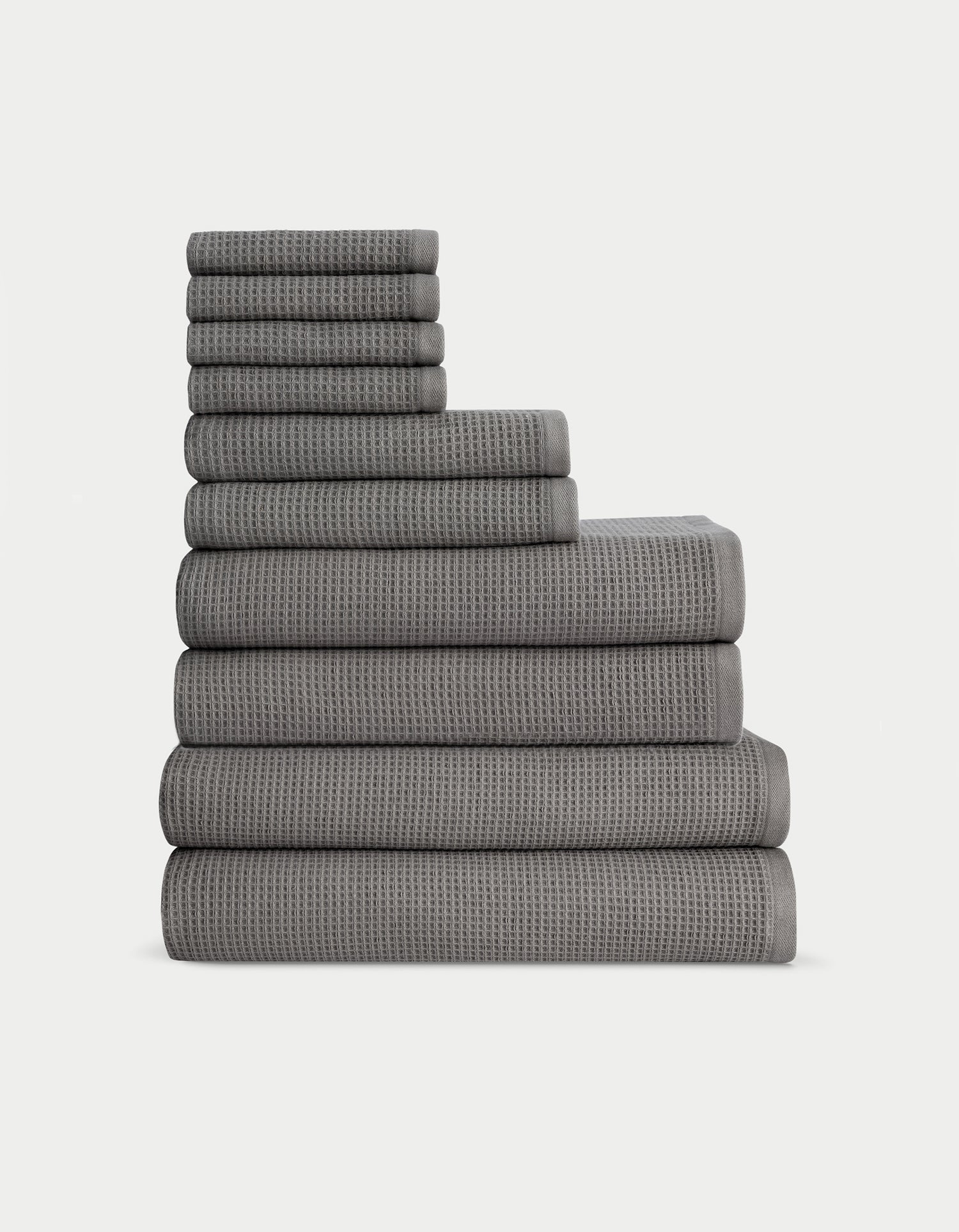 The Complete Waffle Bath Bundle by Cozy Earth features a neatly stacked set of dark gray waffle-textured towels, arranged from largest to smallest, against a plain white background.|Color:Charcoal