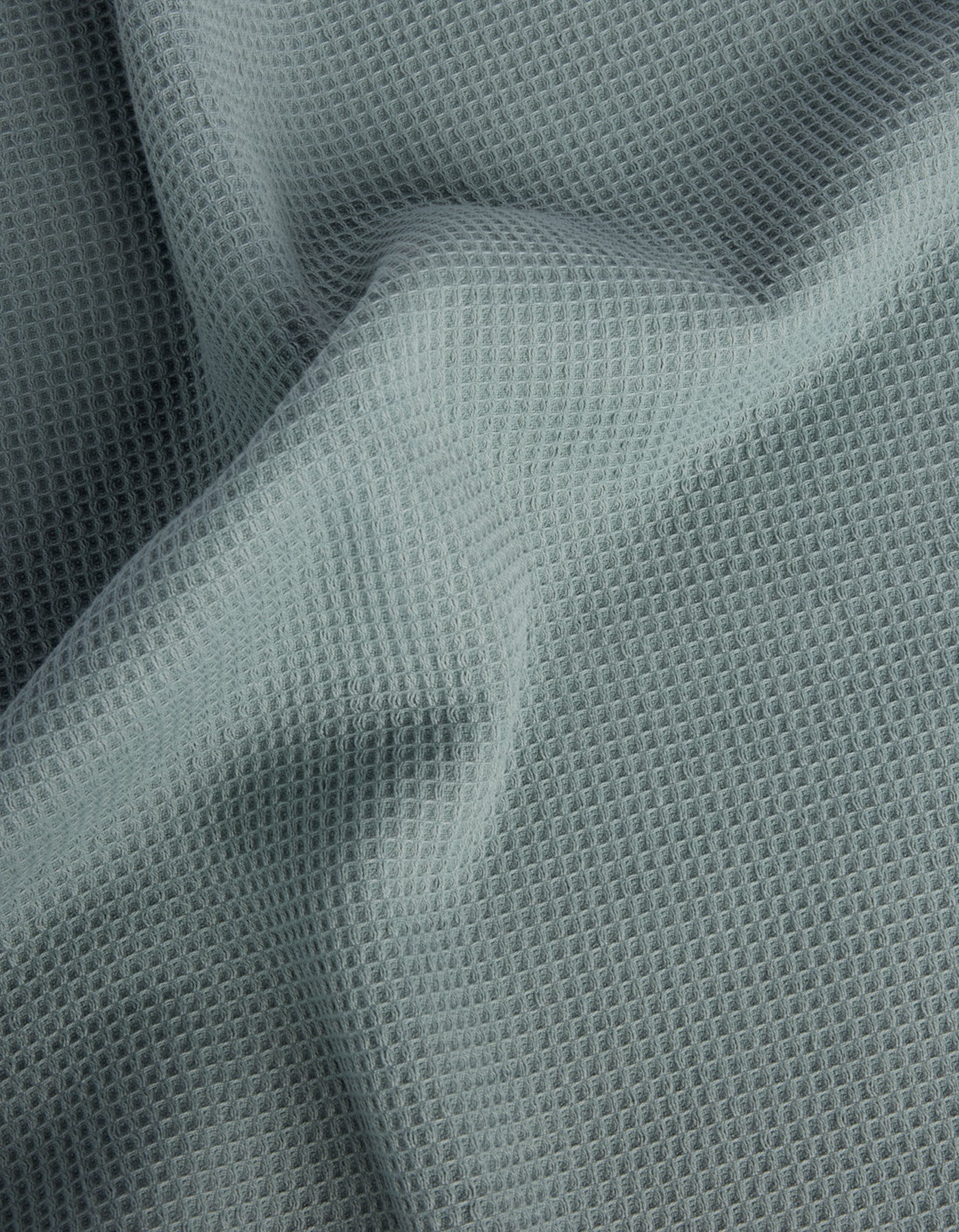 Cozy Earth’s Waffle Bath Sheets feature light gray fabric with a waffle-like texture and a grid of small, raised squares. The soft material drapes gently, creating subtle folds and shadows on the surface. 