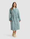 A woman with long brown hair stands facing forward in a light blue Cozy Earth Waffle Bath Robe with a belt and two front pockets, paired with beige slippers. The background is a plain light gray. |Color:Brook