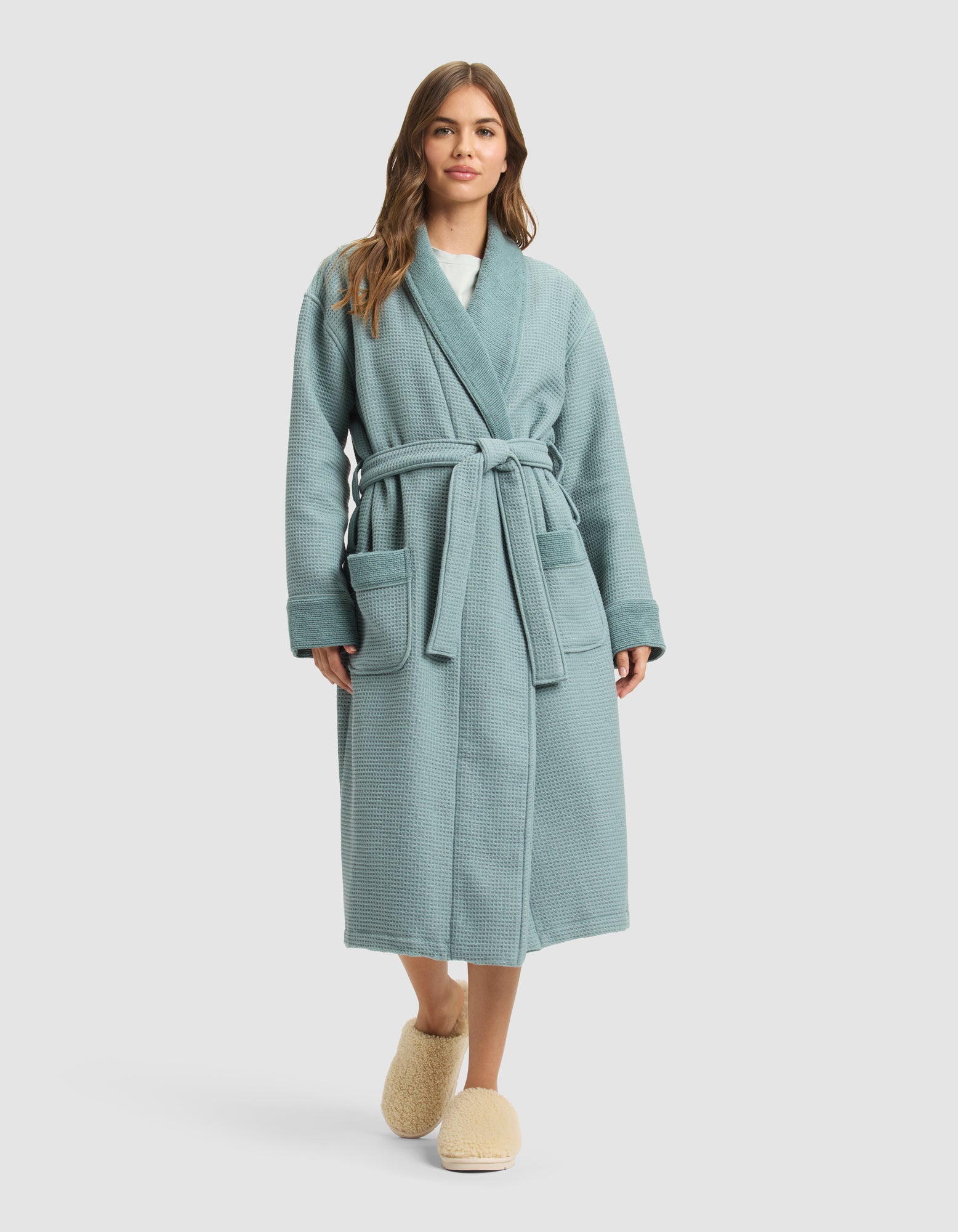 A woman with long brown hair stands facing forward in a light blue Cozy Earth Waffle Bath Robe with a belt and two front pockets, paired with beige slippers. The background is a plain light gray. 