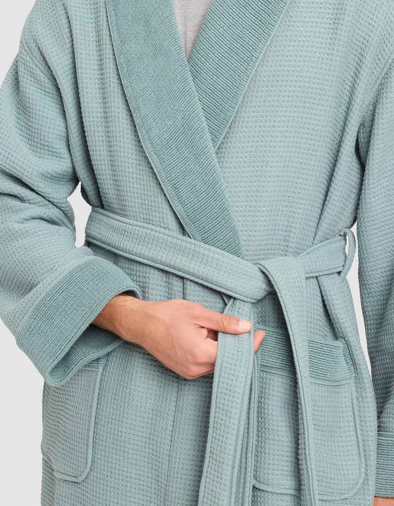 A person is tying the belt of a Cozy Earth Waffle Bath Robe in light blue, holding the belt with their left hand and adjusting it with their right. The textured robe is visible from the shoulders down.
