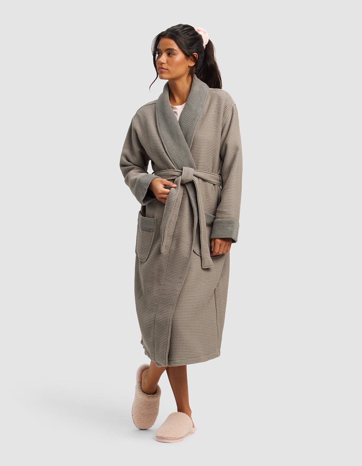 A woman with dark hair tied back wears the Cozy Earth Waffle Bath Robe—grey, textured, with large pockets and a tie belt—and pink fuzzy slippers, standing hands-in-pockets against a plain light background.