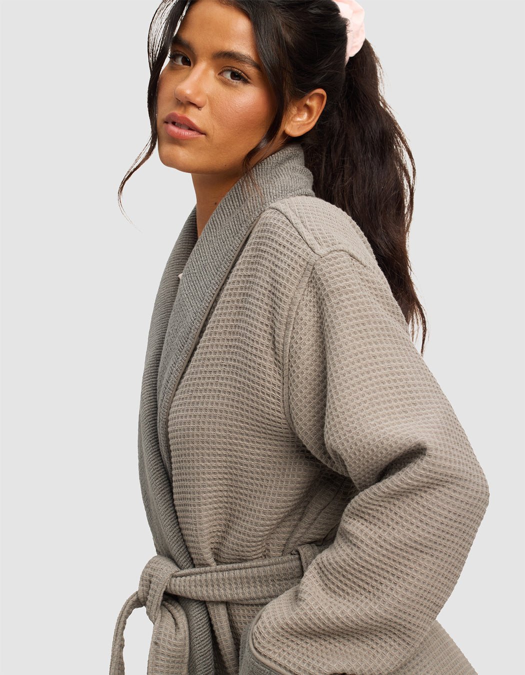 A woman with long dark hair in a ponytail, wearing a light pink scrunchie and the Cozy Earth Waffle Bath Robe with belt, poses against a plain light gray background.|Color:Charcoal