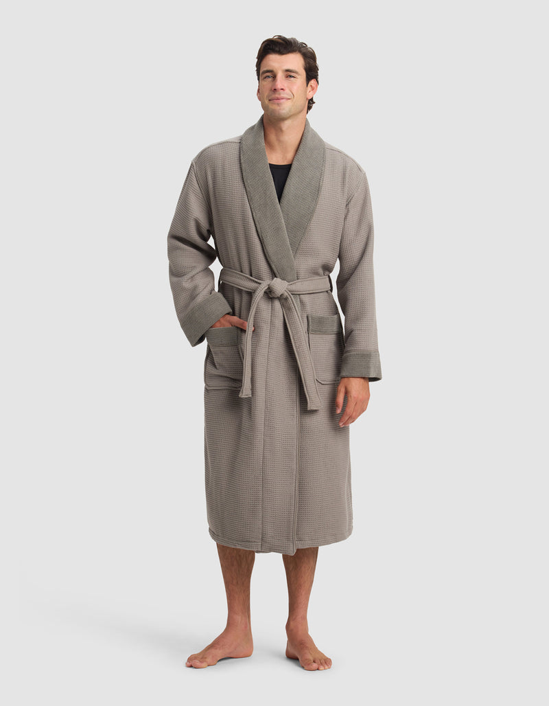A man stands barefoot in a Cozy Earth Waffle Bath Robe—light brown with a darker shawl collar, front pockets, and a tied belt—worn over a black shirt. The background is plain and light-colored.
