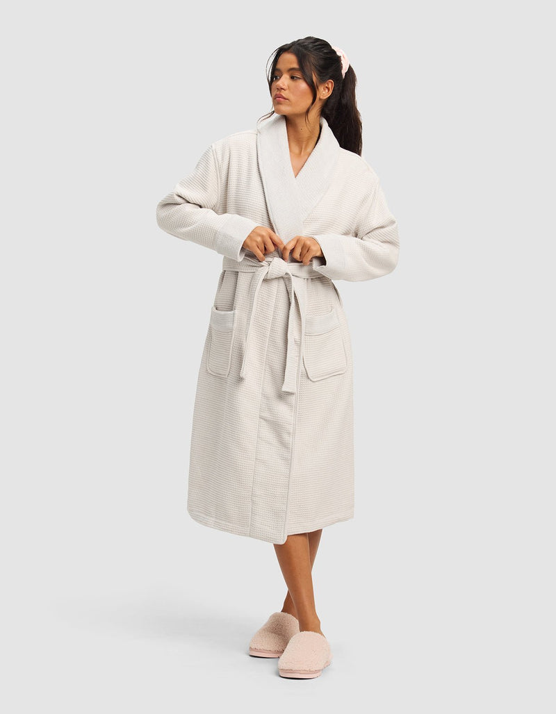 A woman models the Cozy Earth Waffle Bath Robe, featuring a belted waist and large front pockets, paired with fluffy pink slippers. She stands against a plain light background, gazing to the side.