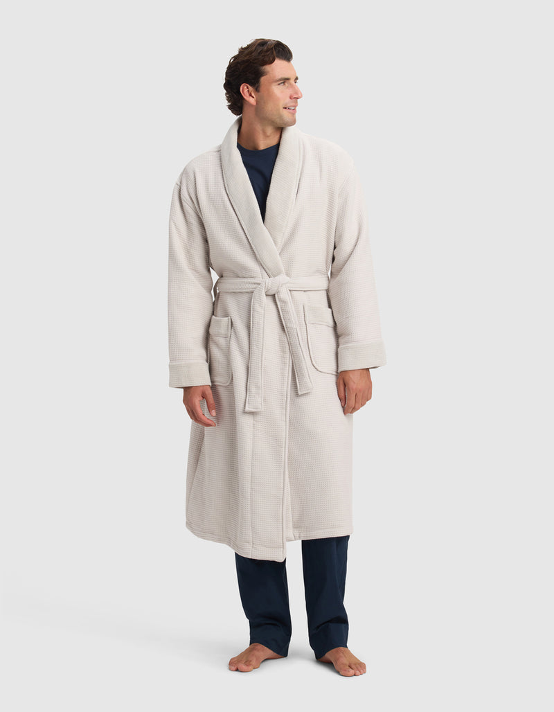 A person wearing a Cozy Earth Waffle Bath Robe, tied at the waist over dark pants and a dark shirt, stands barefoot on a white background, looking off to the side and smiling.