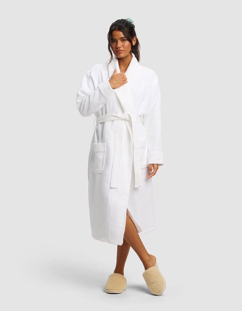 A woman stands against a plain background wearing the Cozy Earth Waffle Bath Robe tied at the waist and beige slippers. Her hair is tied back, with one hand near her collar and one at her side.