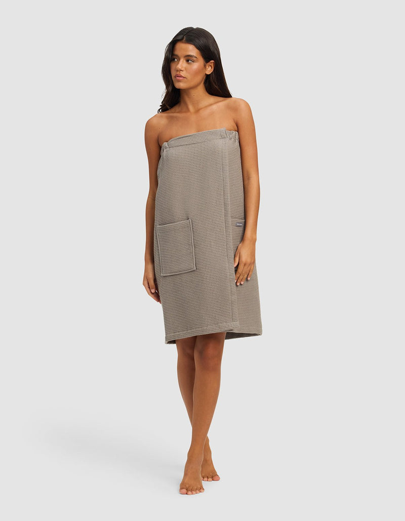 A woman stands barefoot against a plain background, wearing the Cozy Earth Waffle Bath Wrap in grey, featuring a waffle texture, front pocket, and chest-to-knee coverage.