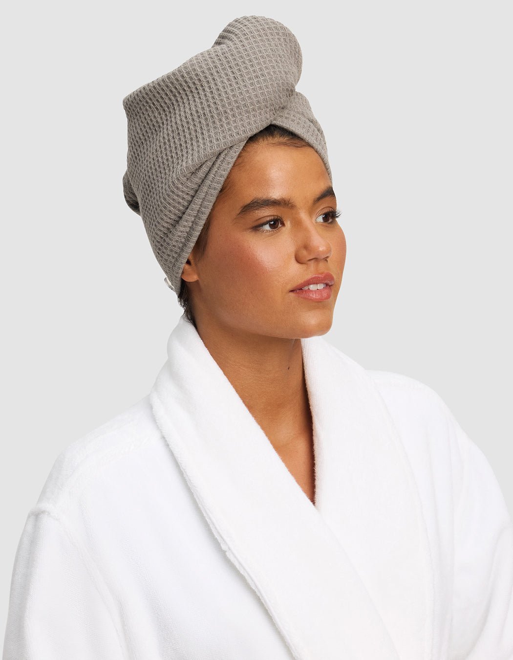 A person wears a white bathrobe and a Cozy Earth Waffle Hair Towel in light gray, wrapped around their head, looking slightly to the side with a neutral expression against a plain light gray background. 