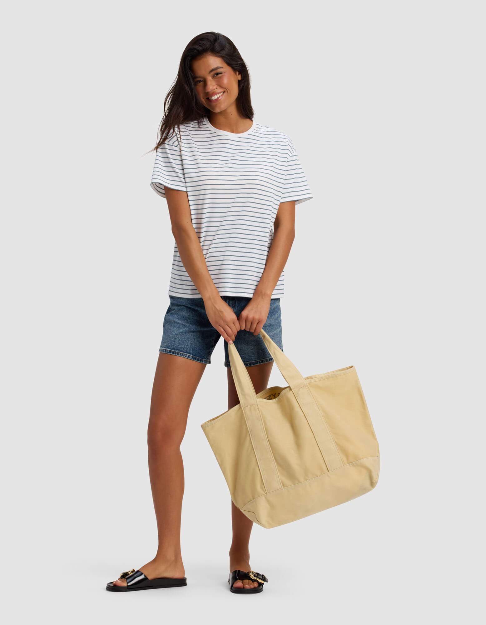 A woman stands smiling against a light gray background, holding the Cozy Earth Waxed Canvas Tote with both hands, dressed in a white and navy striped t-shirt, denim shorts, and black sandals.|Color:Natural