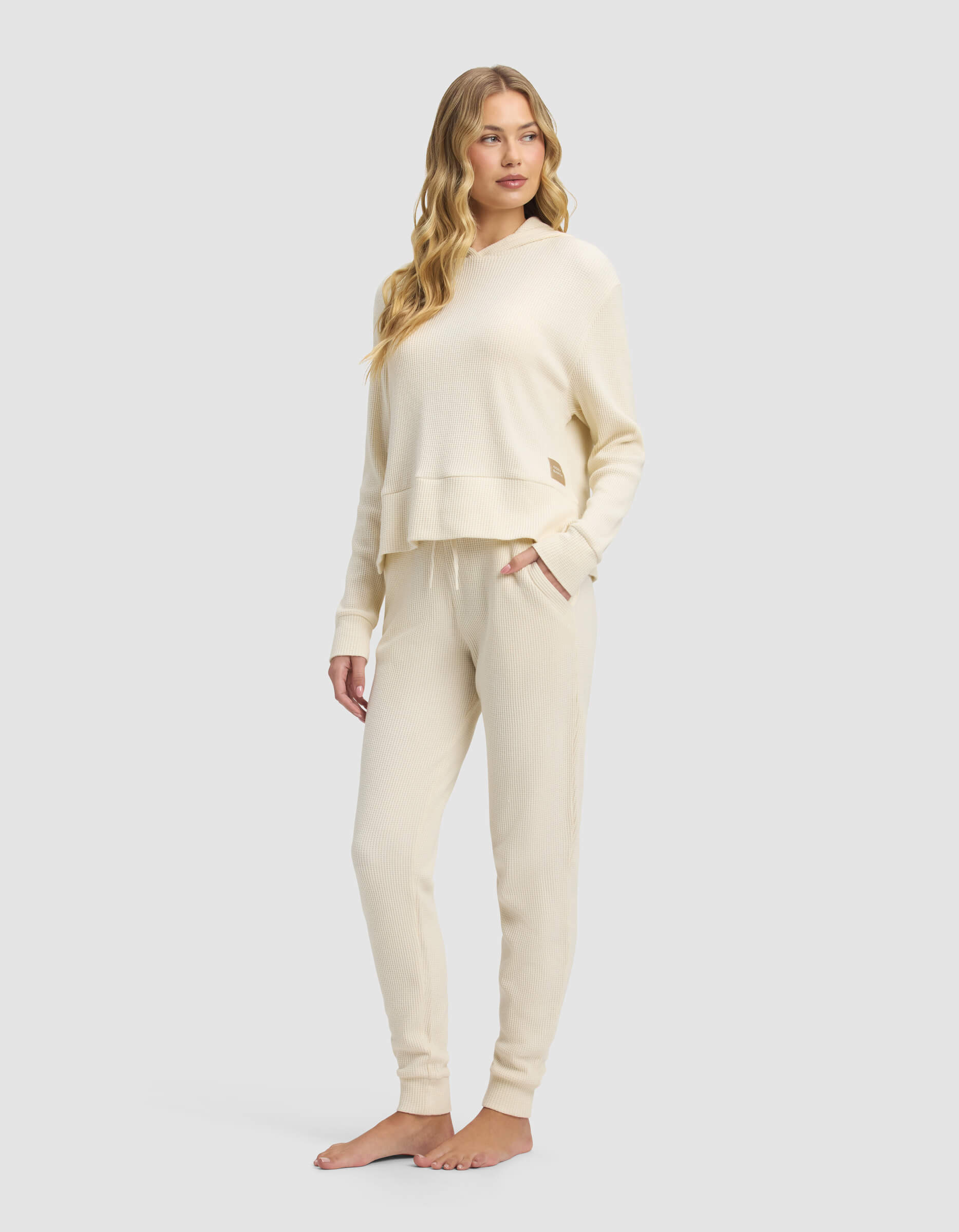 A woman with long, wavy blonde hair wears the Cozy Earth Women's Blake Waffle Hoodie & Jogger Set in cream. She stands barefoot against a plain light gray background.