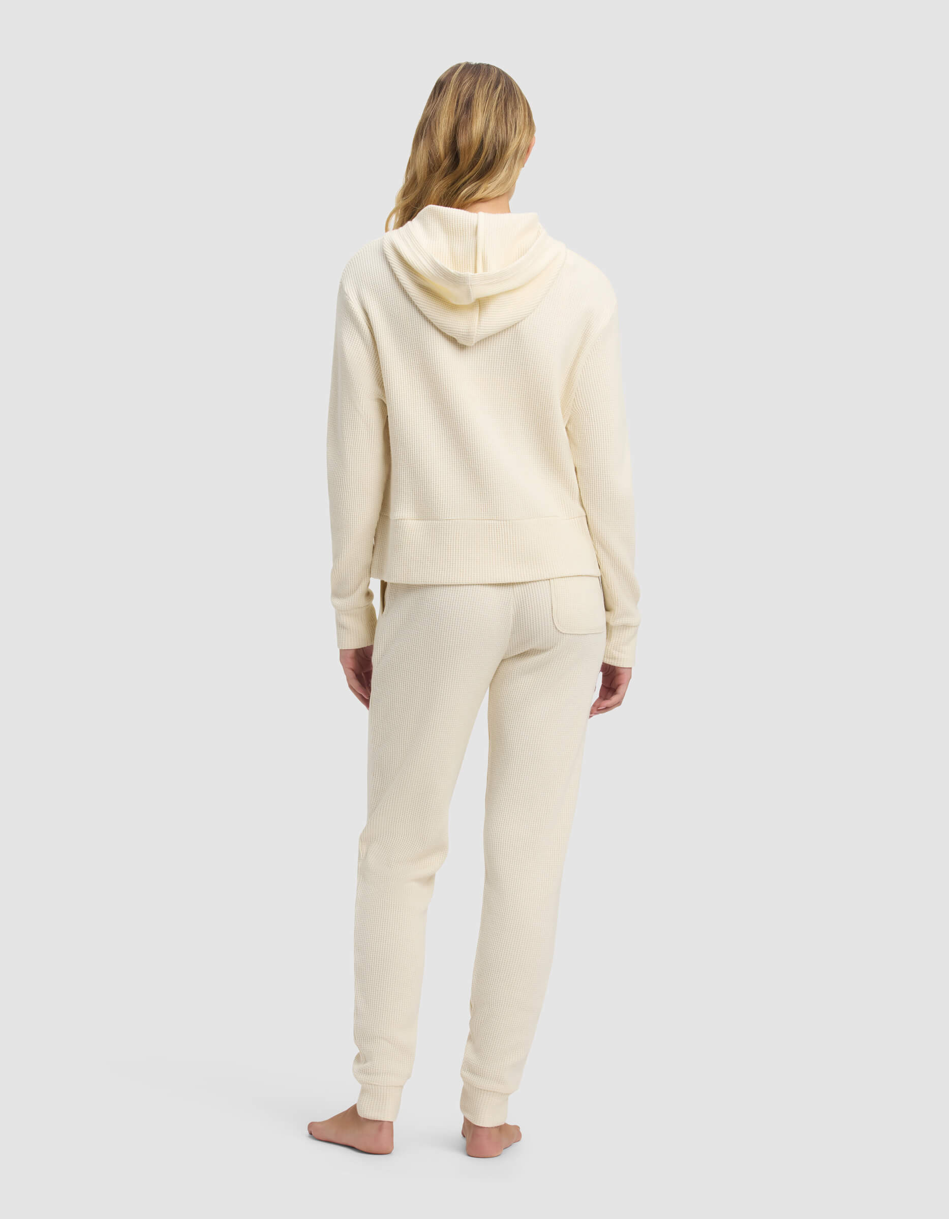 A woman with long blonde hair stands barefoot, facing away, wearing the Cozy Earth Women's Blake Waffle Hoodie & Jogger Set in cream against a plain light gray background.|Color:Coconut