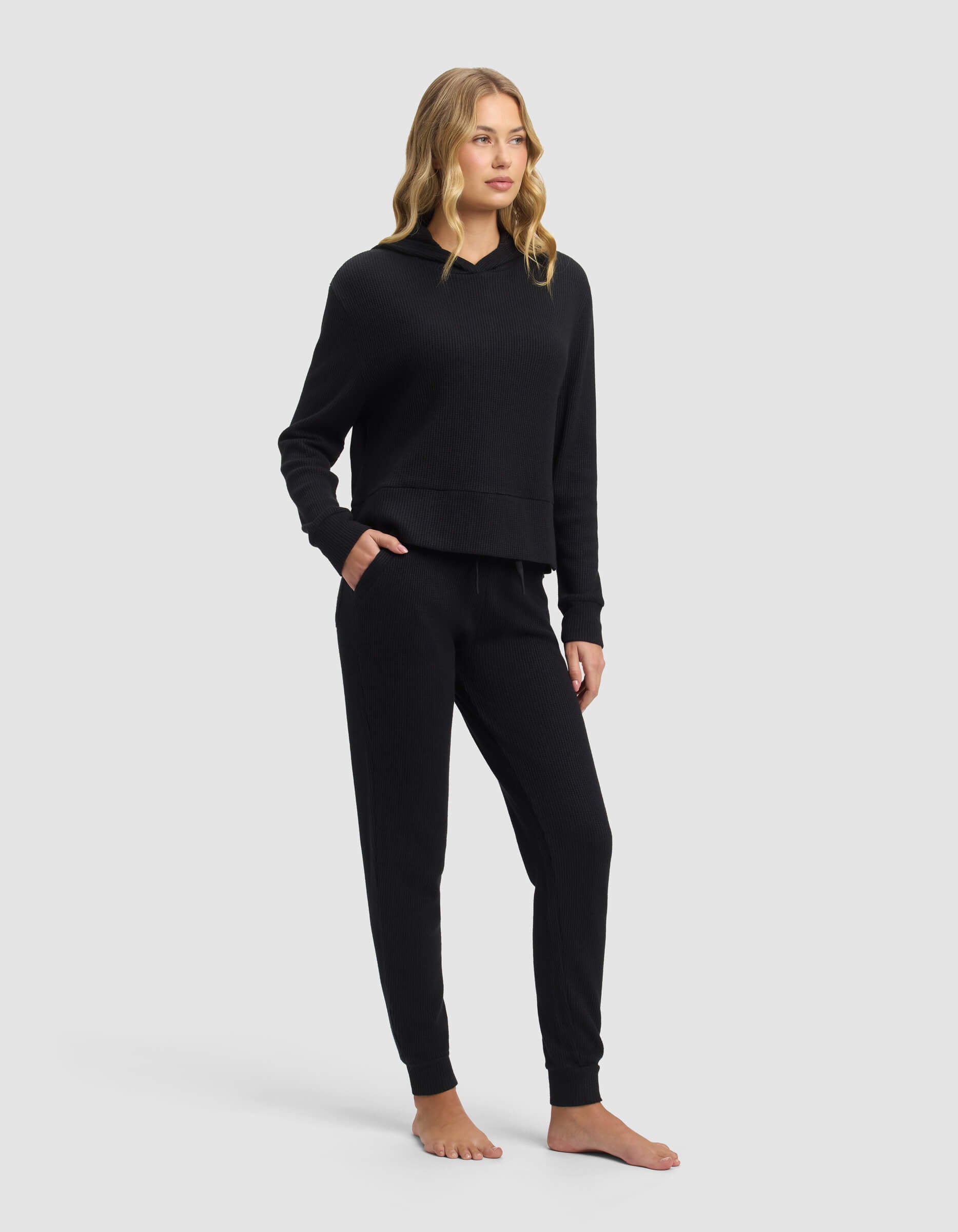 A woman with long blonde hair stands barefoot, wearing the Cozy Earth Women's Blake Waffle Hoodie & Jogger Set in black. She poses with one hand in her pocket, glancing to the side against a plain light gray background.