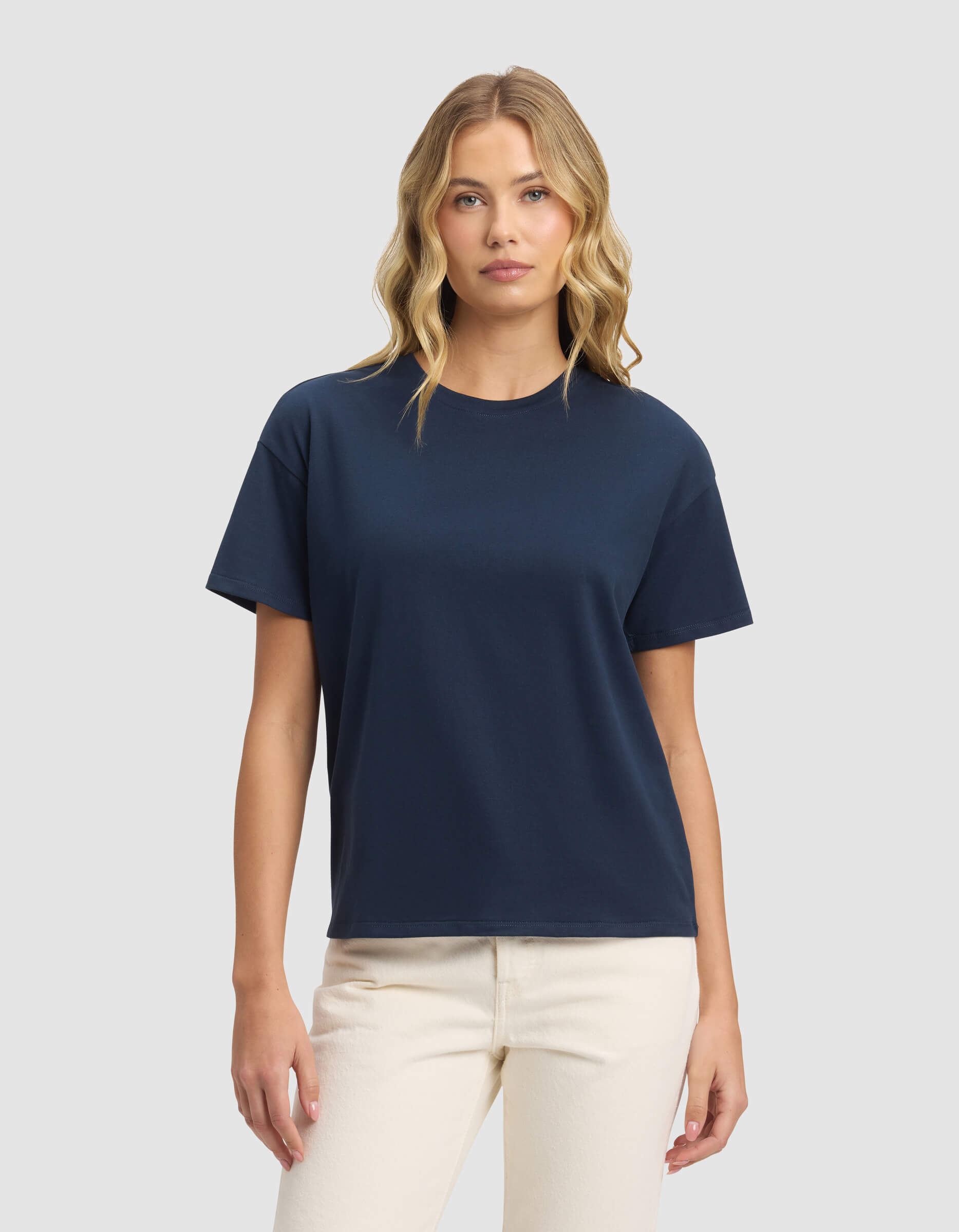 A woman with wavy blonde hair wears the Cozy Earth Women's All Day Tee in navy blue and light beige pants, standing against a light gray background and facing forward with a neutral expression.