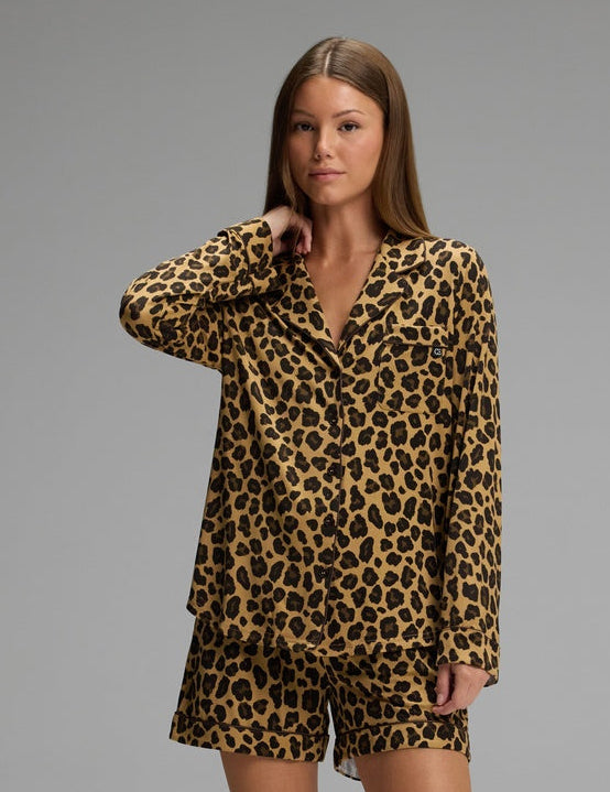 A woman with straight brown hair and a neutral expression stands barefoot against a gray background wearing the Cozy Earth Women’s Bamboo Stretch-Knit Long Sleeve & Short Pajama Set in Leopard Print in Onyx.