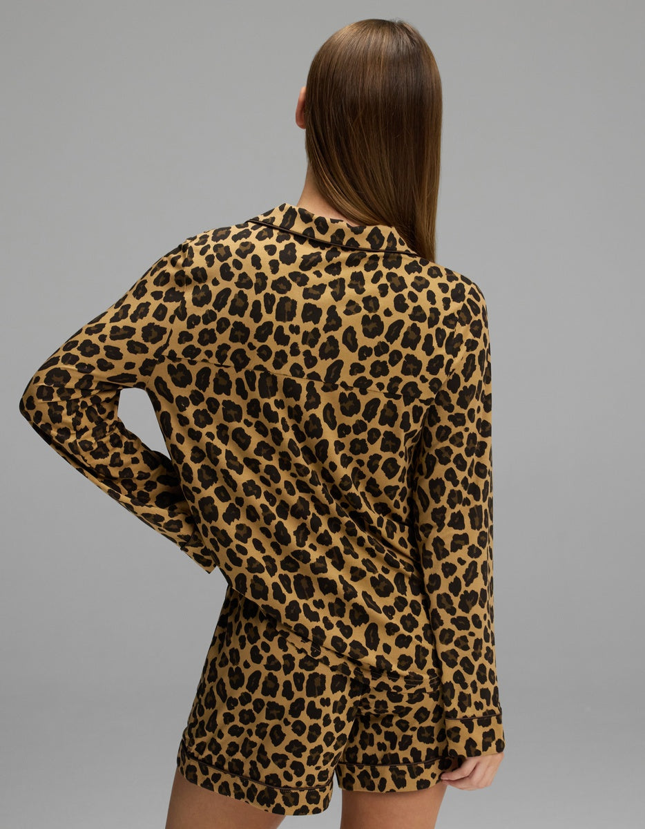 A person with straight brown hair models the Women’s Bamboo Stretch-Knit Long Sleeve & Short Pajama Set by Cozy Earth, standing with one hand on their hip and facing away from the camera against a gray background.