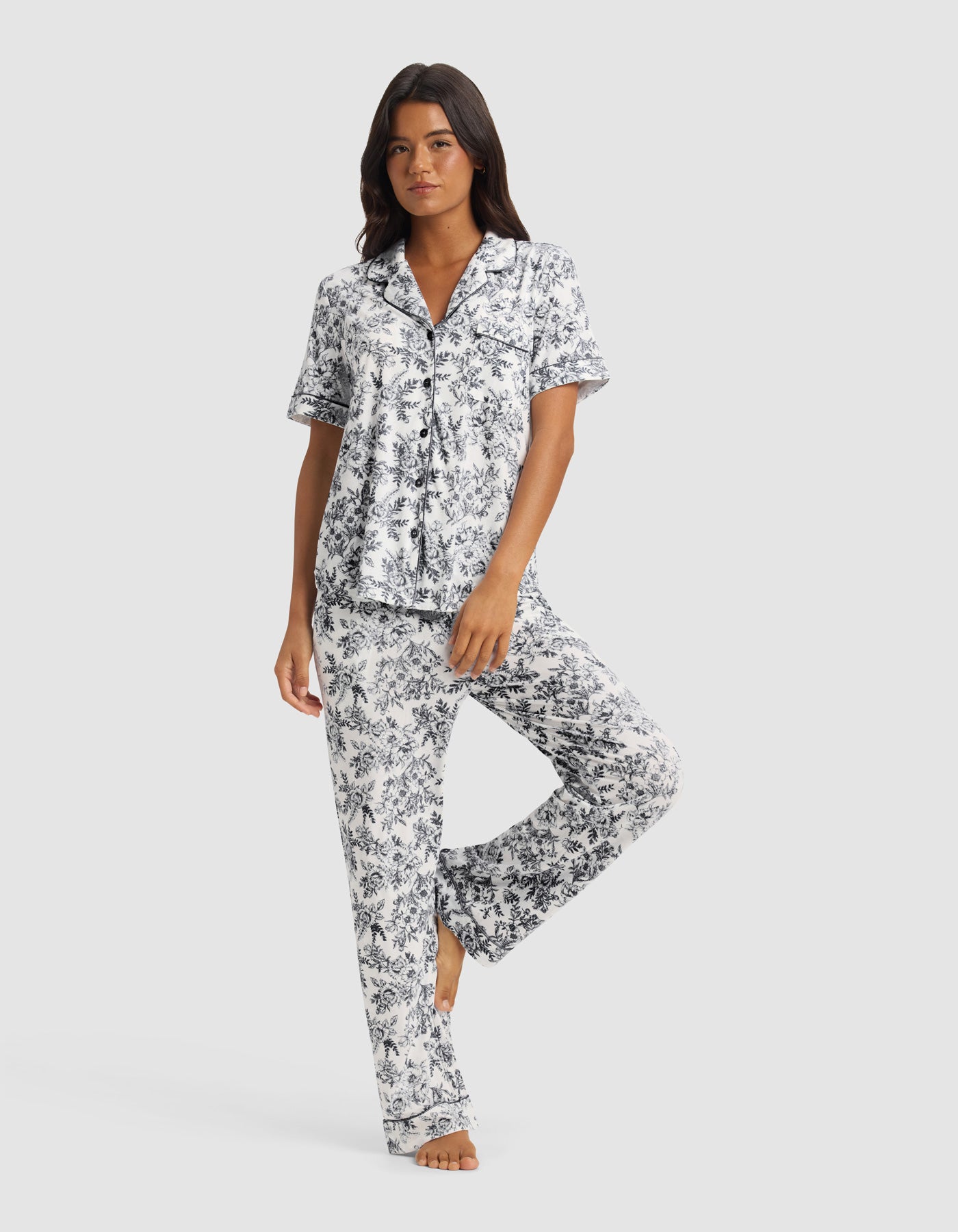 A woman wears the Cozy Earth Women’s Bamboo Stretch-Knit Short Sleeve & Pant Pajama Set in Jet Black Toile, standing barefoot on one leg with the other slightly bent against a plain light background.