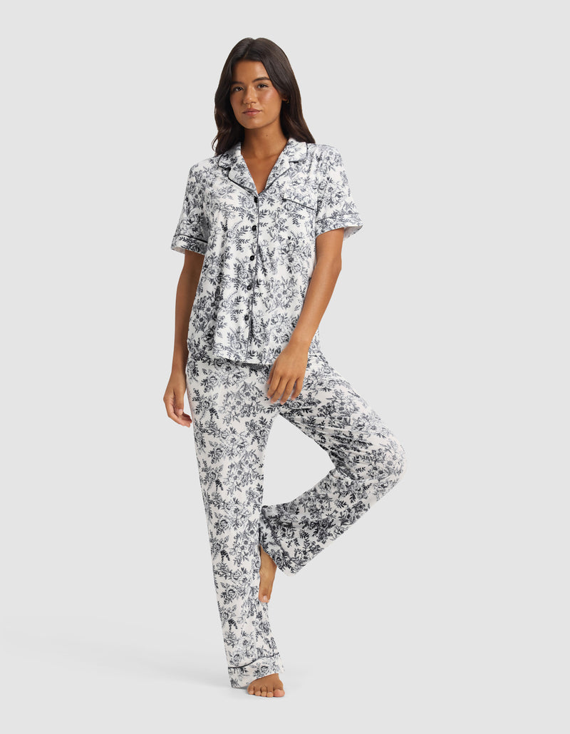 A woman wears the Cozy Earth Women’s Bamboo Stretch-Knit Short Sleeve & Pant Pajama Set in Jet Black Toile, standing barefoot on one leg with the other slightly bent against a plain light background.