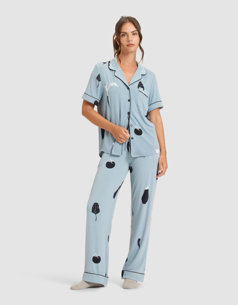 A woman wears HIDE's Women's Bamboo Stretch-Knit Pajama Pant in light blue with black and white fruit and leaf print, paired with gray socks. The set features short sleeves, buttons, a collar, and is shown against a plain white background.