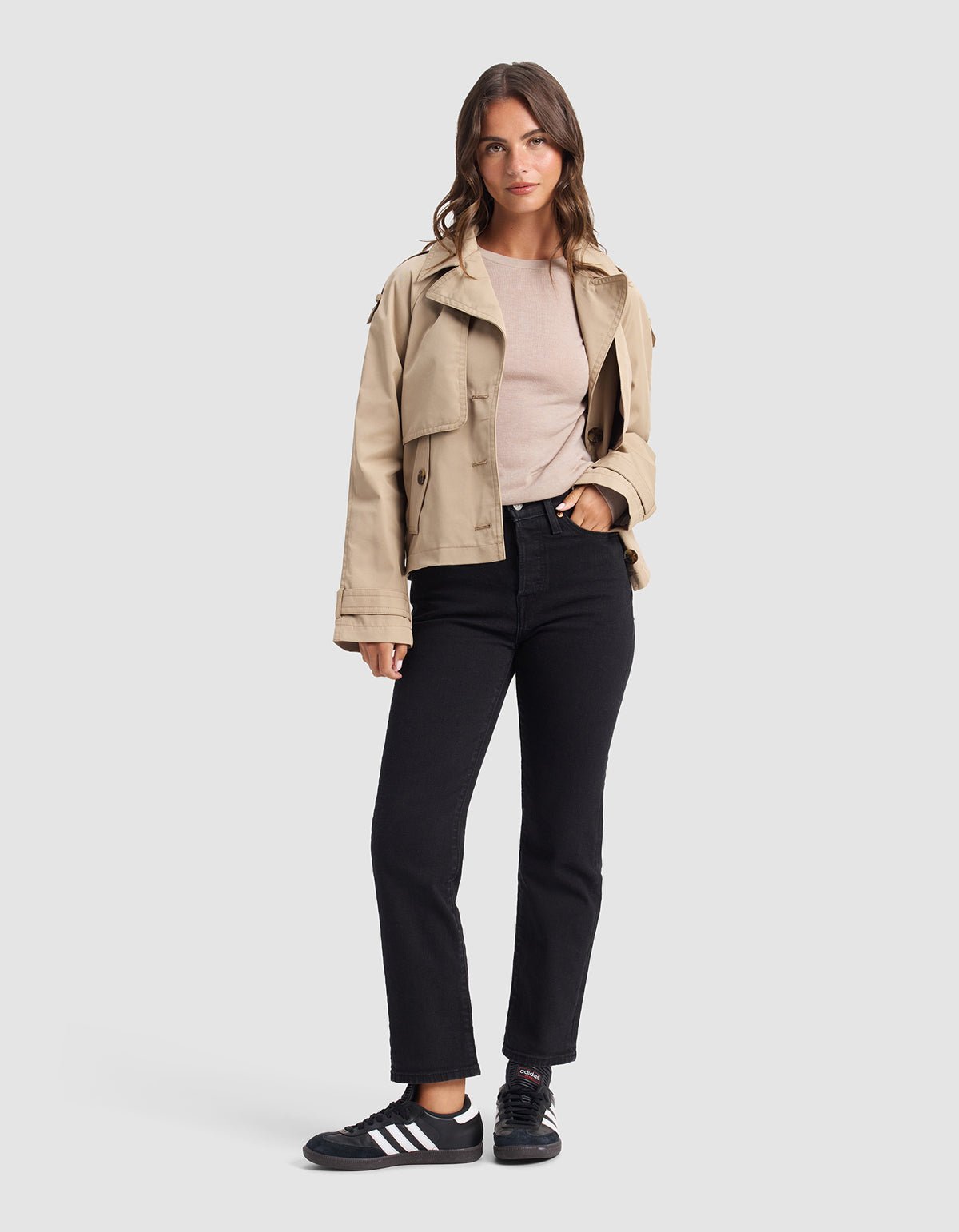 A woman faces forward, wearing the Cozy Earth Women's AirKnit Crewneck Sweater in beige, black jeans, and black Adidas sneakers with white stripes, standing against a plain gray background.