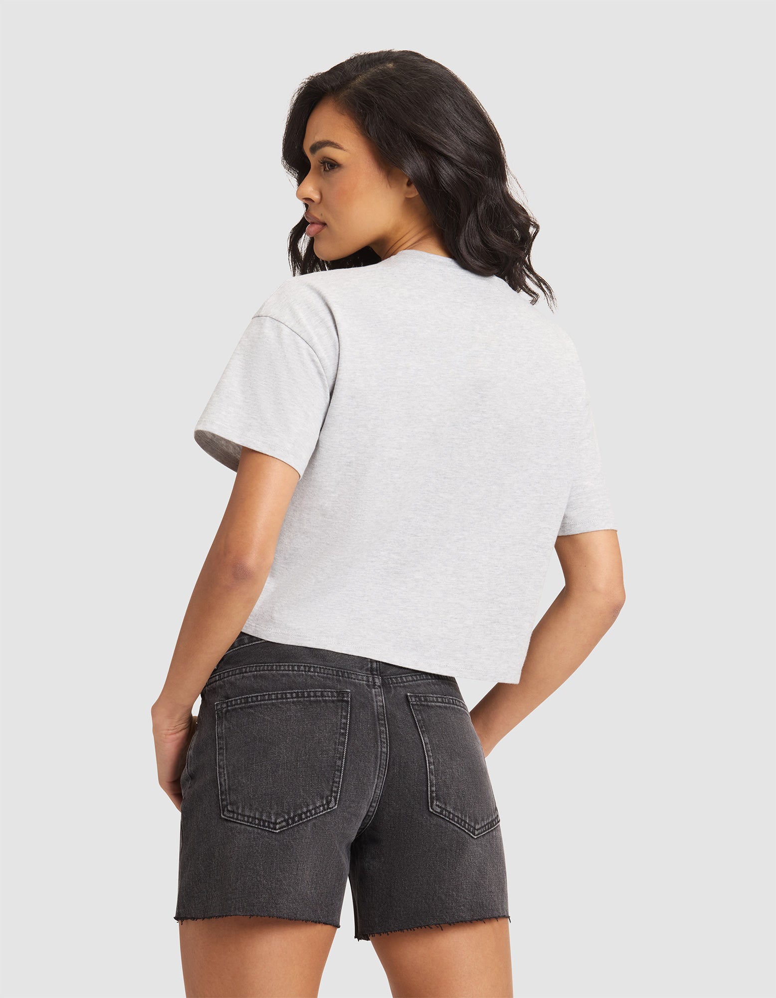 A woman with dark hair stands facing away, wearing the Cozy Earth Women's All Day Cropped Tee in light gray and black denim shorts, set against a plain light background. 