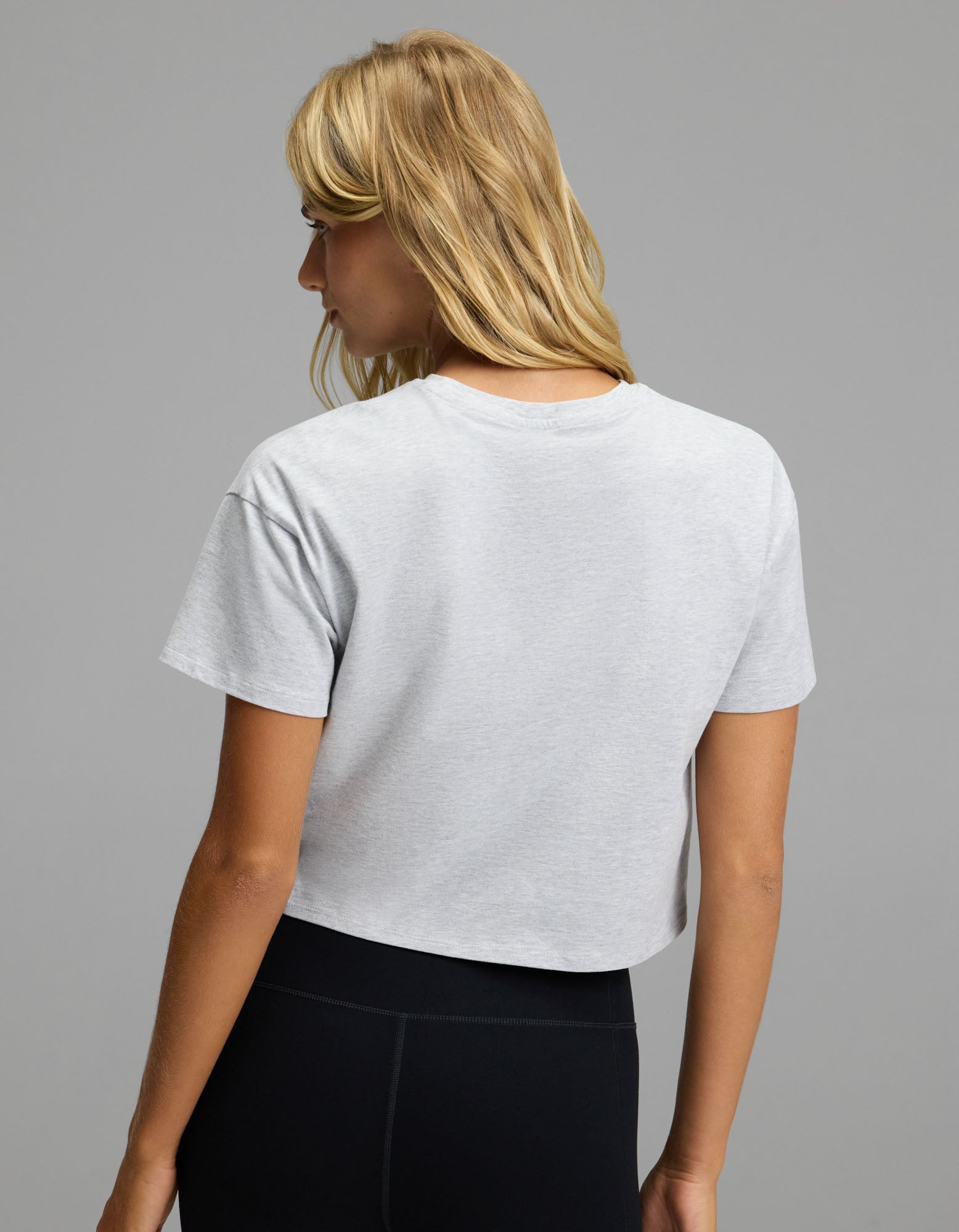 A person with long blonde hair is seen from behind against a plain gray background, wearing the Cozy Earth Women’s All Day Cropped Tee in light gray paired with black leggings.