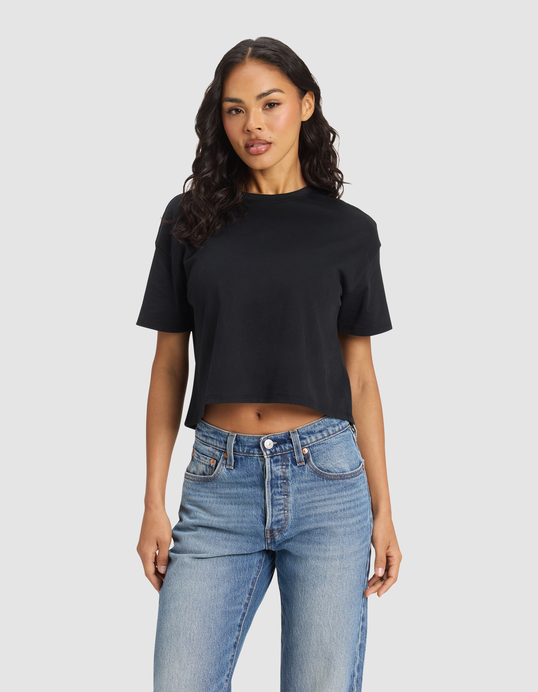 A woman with long, wavy hair wears the black Cozy Earth Women's All Day Cropped Tee and blue jeans, standing against a plain light gray background. 