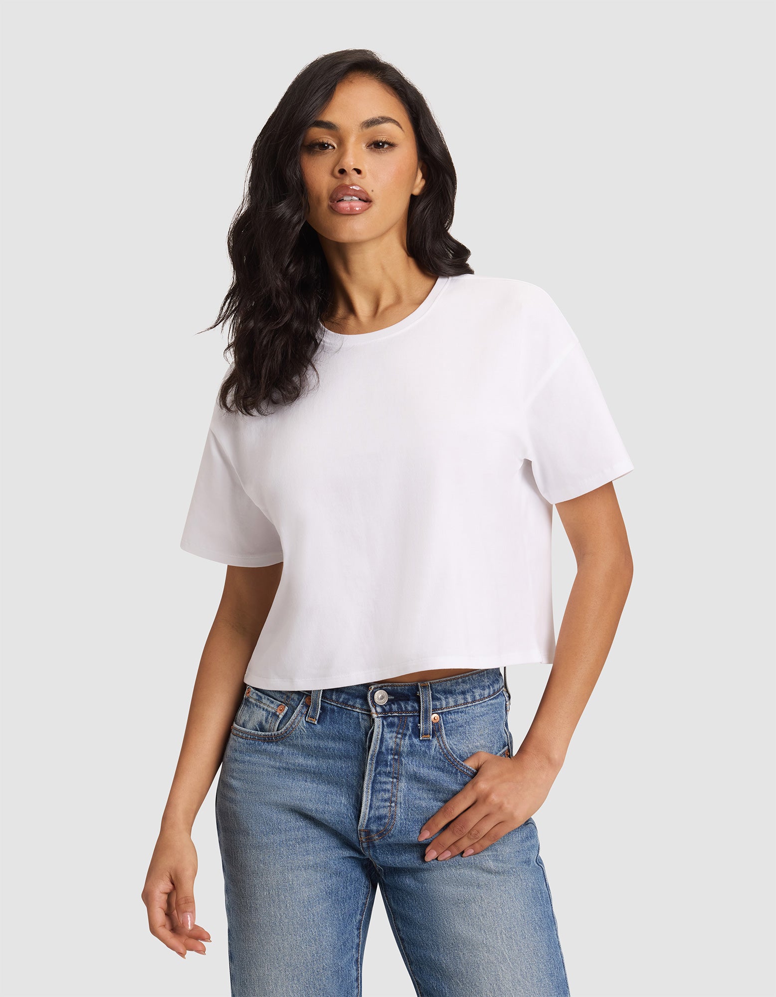 A woman with long dark hair models the Cozy Earth Women's All Day Cropped Tee in white with blue jeans, standing against a light gray background and posing with one hand in her pocket. |Color:White
