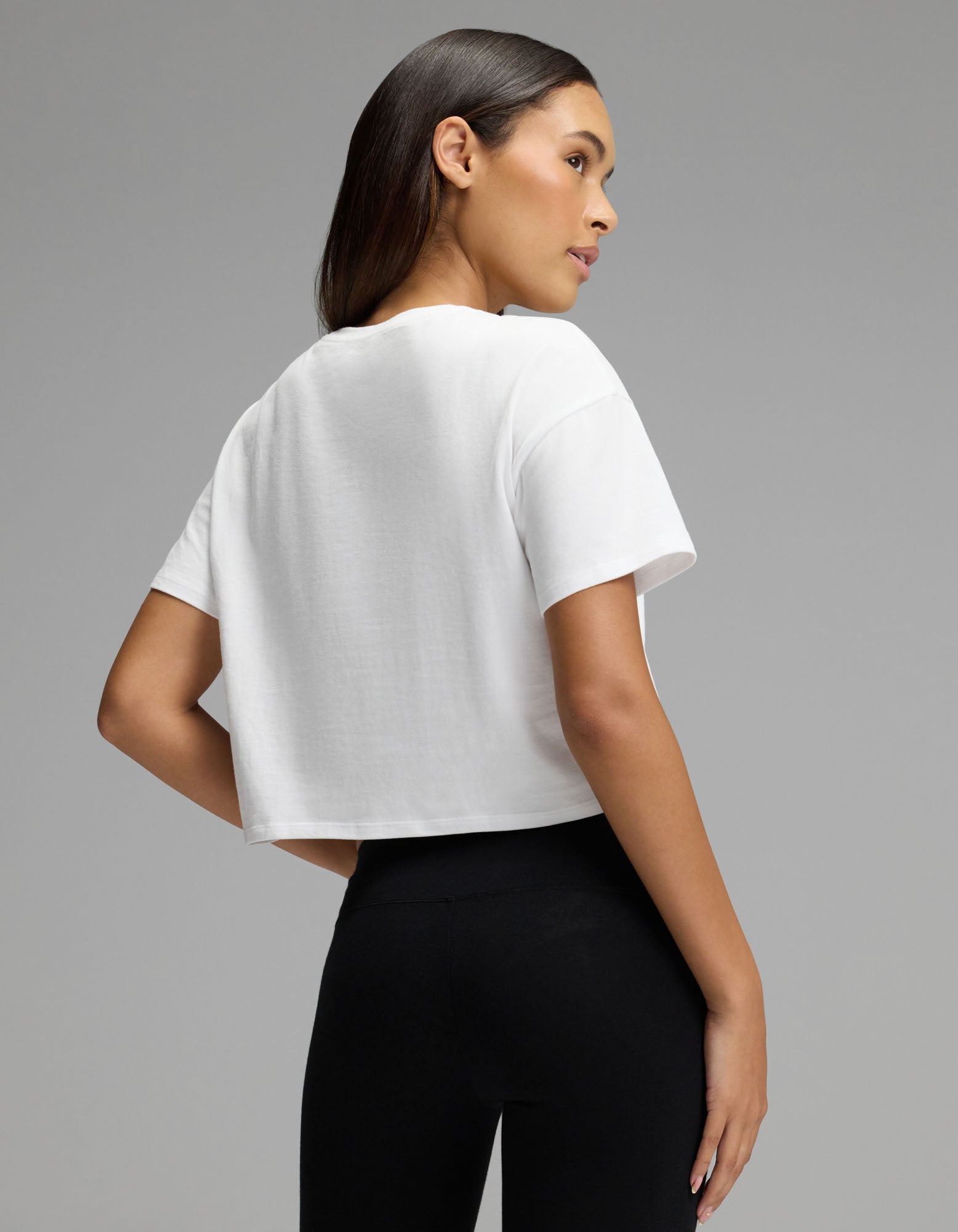 A woman with long dark hair models the Cozy Earth Women’s All Day Cropped Tee in white, paired with black leggings. She stands sideways, looking over her shoulder against a plain gray background.