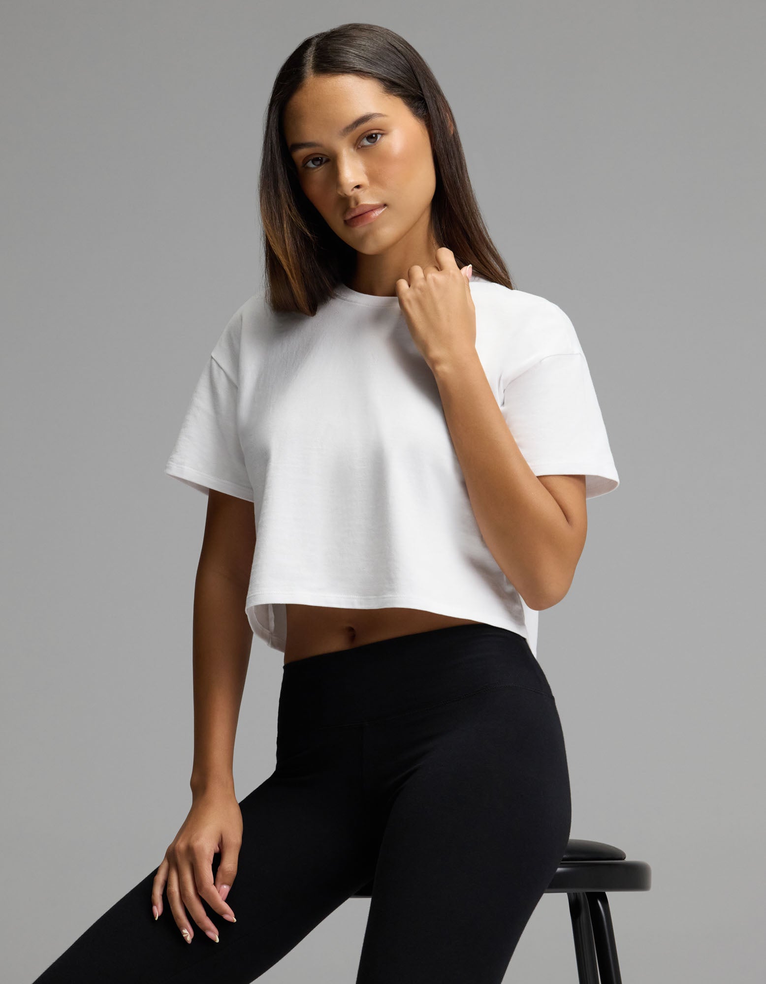 A woman models the Cozy Earth Women’s All Day Cropped Tee in white and black leggings, seated on a stool and looking at the camera with a neutral expression against a plain gray background.|Color:White