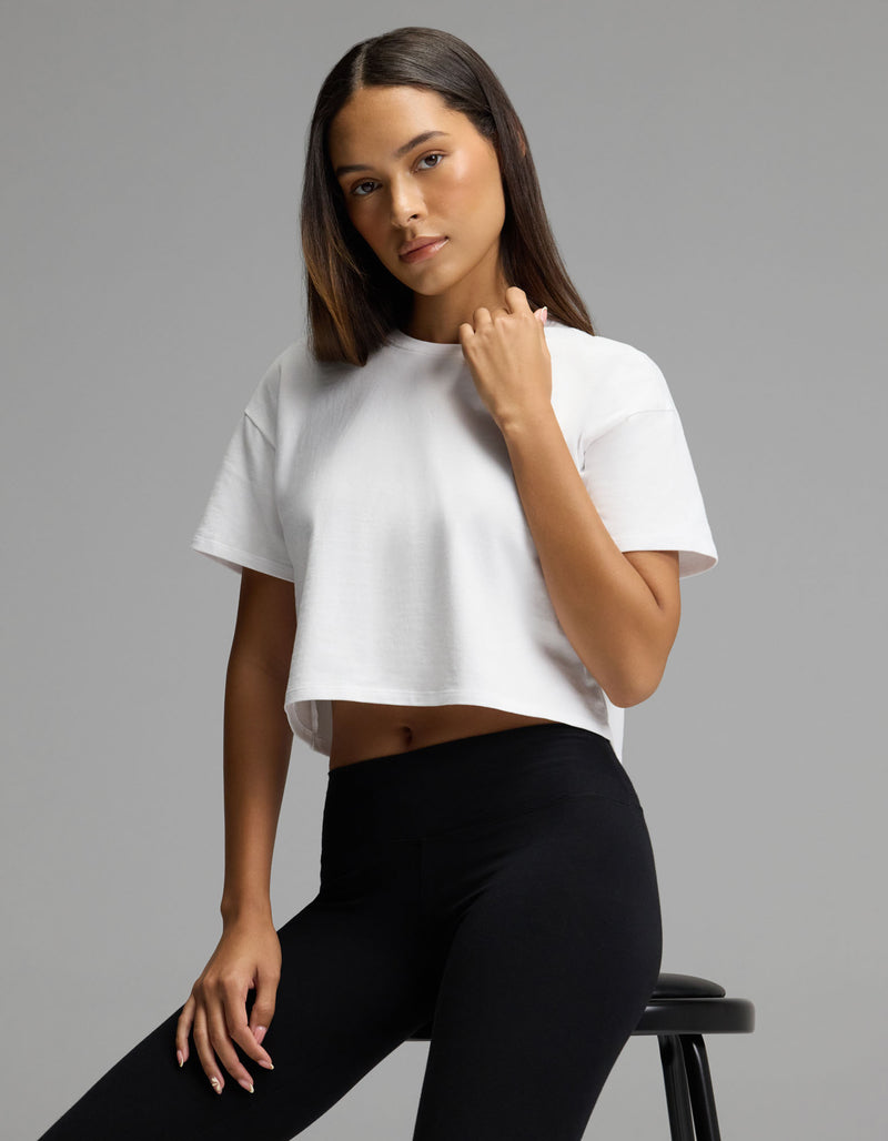 A woman models the Cozy Earth Women’s All Day Cropped Tee in white and black leggings, seated on a stool and looking at the camera with a neutral expression against a plain gray background.