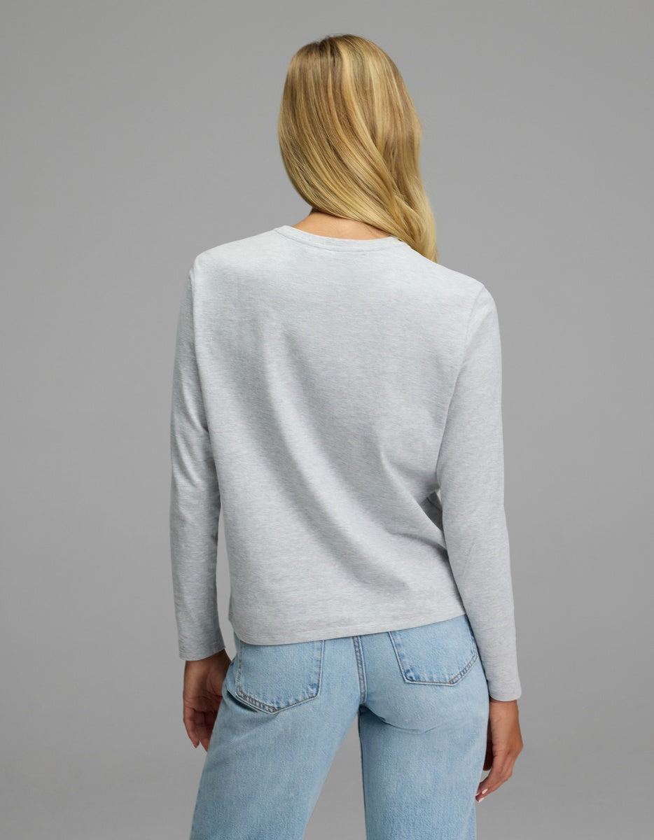 A woman with long blonde hair stands facing away, wearing the Cozy Earth Women’s All Day Long Sleeve Tee in light gray and light blue jeans against a plain gray background.