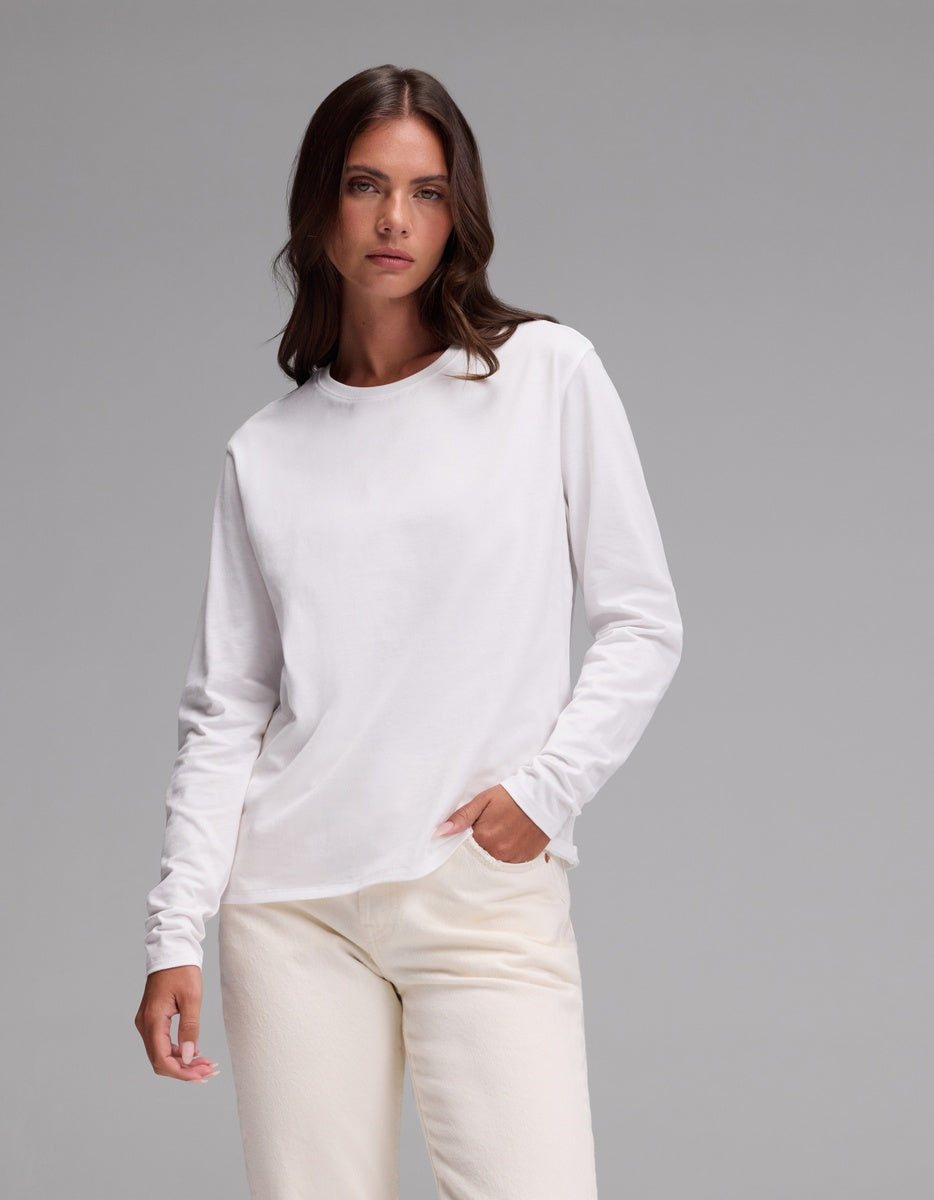 A person with long brown hair wears the Cozy Earth Women’s All Day Long Sleeve Tee in plain white and off-white pants, posing with one hand in their pocket against a plain gray background.|Color:White