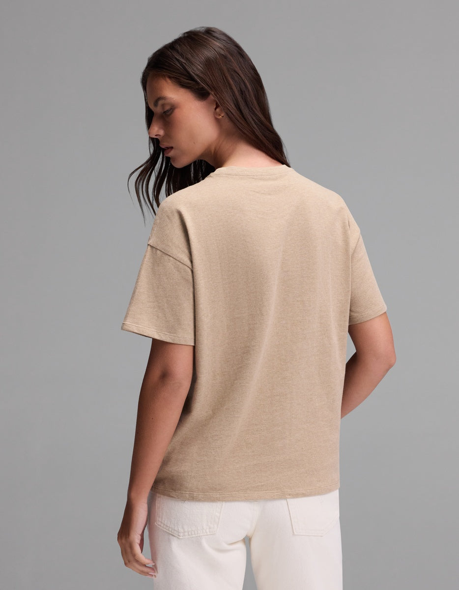 A woman with long brown hair stands facing away, wearing the Cozy Earth Women’s All Day Tee in beige and white pants against a plain gray background.