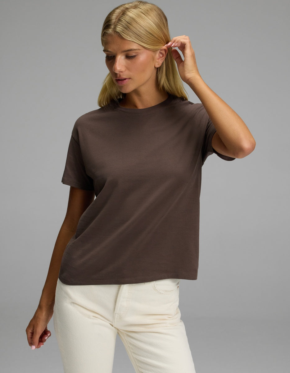 A woman wearing the Cozy Earth Women’s All Day Tee in brown and off-white pants stands against a plain gray background, gently touching her hair and looking down with a neutral expression.|Color:Cacao