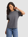A woman with long, wavy hair wears the Cozy Earth Women's All Day Tee in gray and blue jeans, standing against a light gray background while looking down and touching her ear. |Color:Coal Heather